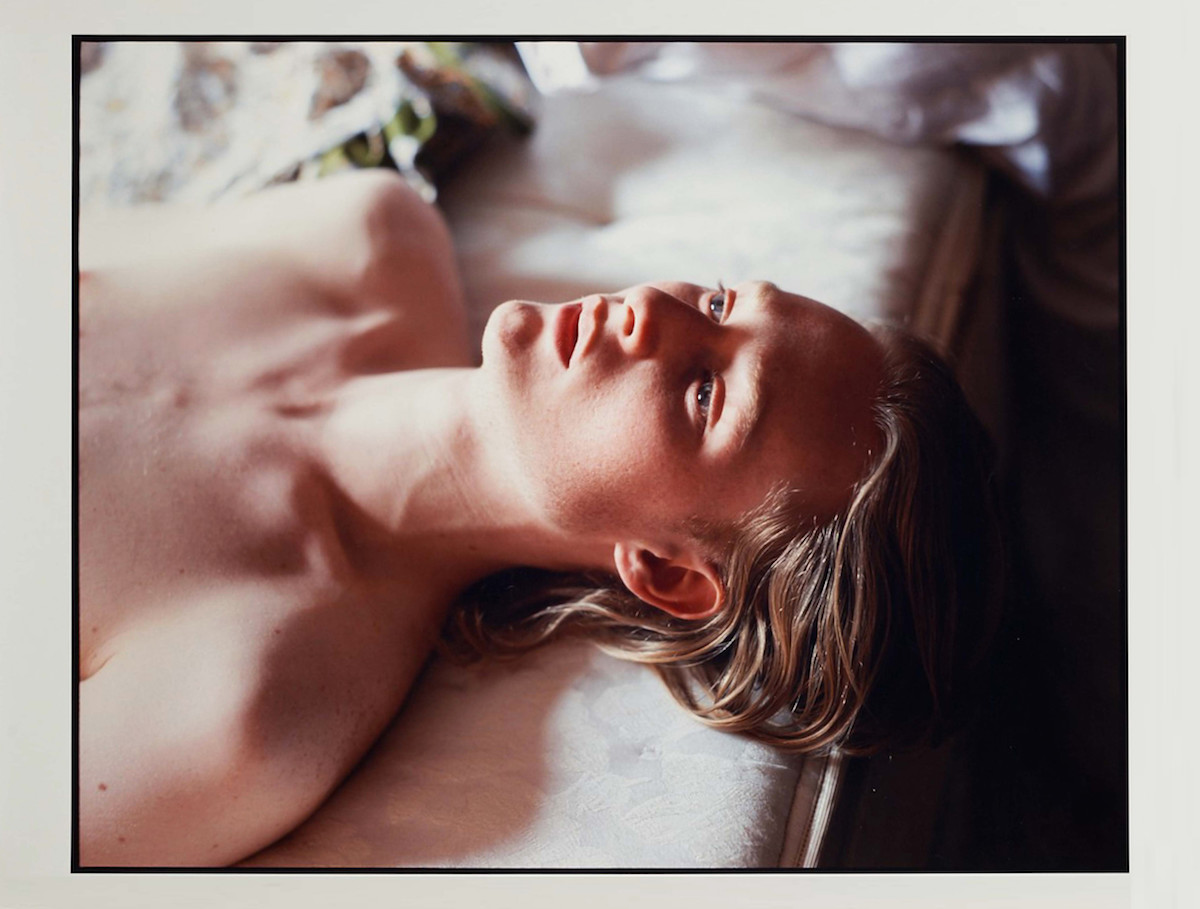 David Armstrong, Koos, 2003, C-print © David Armstrong. Courtesy of the Estate of David Armstrong [A figure lays face up on a white mattress. His expression is languid and his hair falls over the edge of the mattress.]